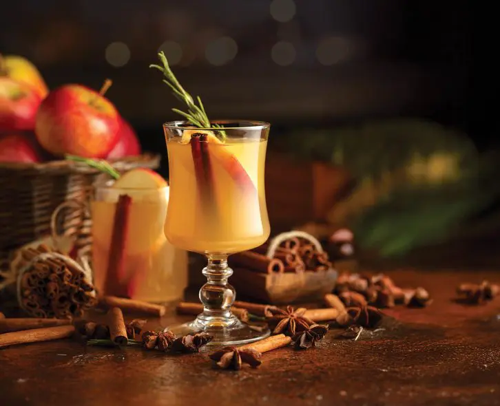 Mulled cider with slice apples, cinnamon, rosemary, and anise stars in glass cups on a brown rustic table. Holiday traditional hot drink. On a dark background kitchen utensils and burning candles.