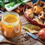 High angle view of healthy homemade bone broth in an open airtight glass container shot on rustic wooden table. Ingredients for cooking bone broth are all around the container. High resolution 42Mp studio digital capture taken with Sony A7rII and Sony FE 90mm f2.8 macro G OSS lens