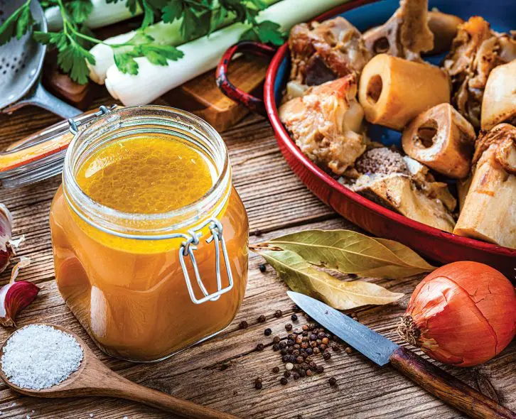 High angle view of healthy homemade bone broth in an open airtight glass container shot on rustic wooden table. Ingredients for cooking bone broth are all around the container. High resolution 42Mp studio digital capture taken with Sony A7rII and Sony FE 90mm f2.8 macro G OSS lens