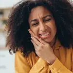 Young African American woman suffering from strong toothache touching her cheek because of acute pain at home