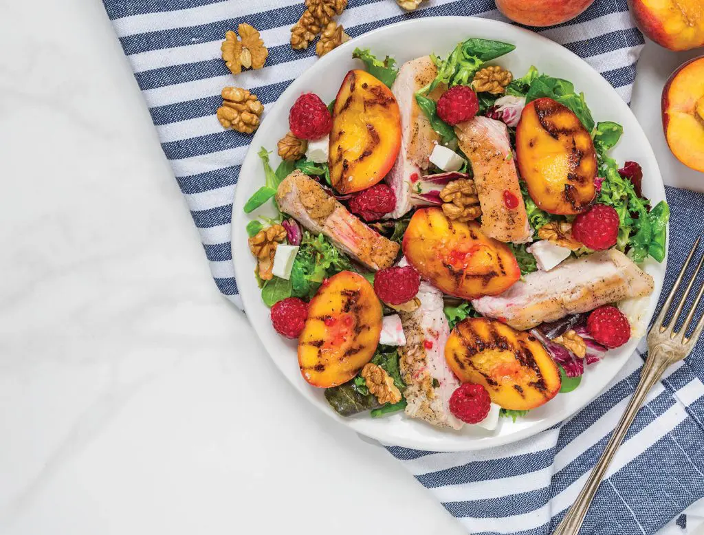 Grilled chicken fillet salad with grilled peaches, raspberries, walnuts and mixed greens on a plate with napkin and fork on white marble background. Top view. Healthy diet food. Summer dinner.