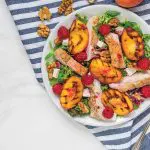 Grilled chicken fillet salad with grilled peaches, raspberries, walnuts and mixed greens on a plate with napkin and fork on white marble background. Top view. Healthy diet food. Summer dinner.