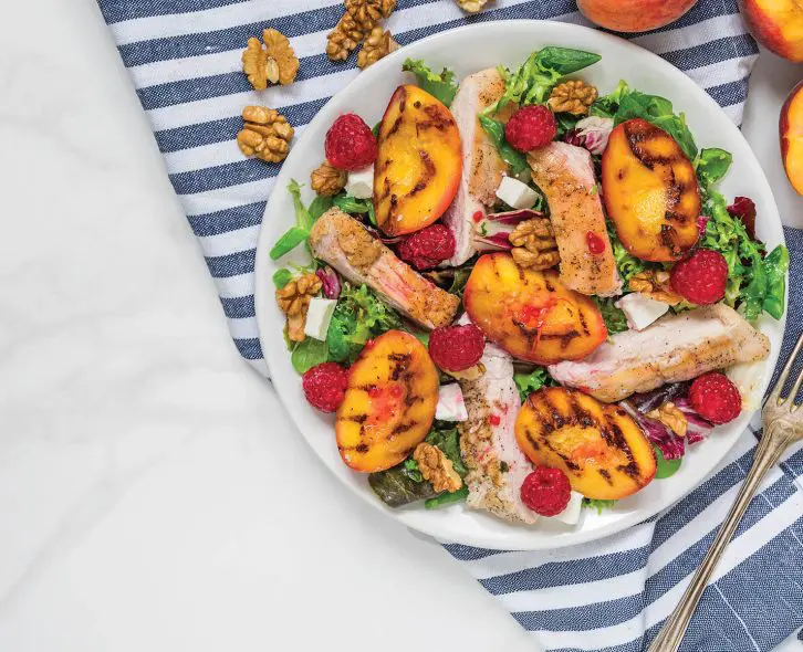 Grilled chicken fillet salad with grilled peaches, raspberries, walnuts and mixed greens on a plate with napkin and fork on white marble background. Top view. Healthy diet food. Summer dinner.