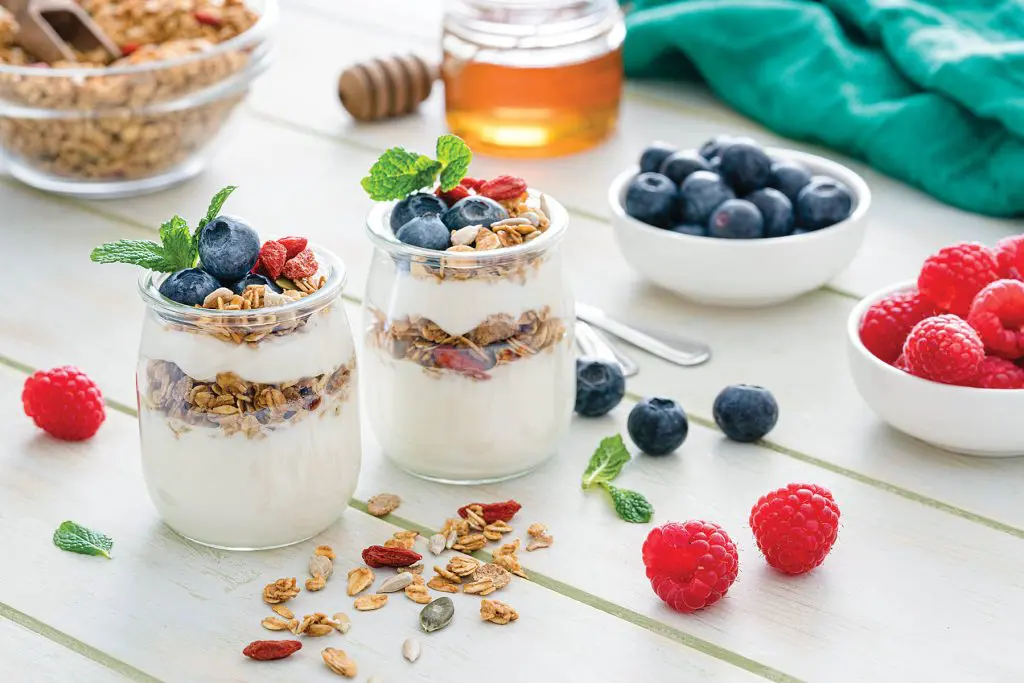 Protein-Packed Yogurt Bowl. Healthy eating: two glass containers with homemade yogurt and granola shot on breakfast table. Two spoons are beside the yogurt containers. Some berries complete the composition. The composition is at the left of an horizontal frame leaving useful copy space for text and/or logo at the right. Predominant color is green. High resolution 42Mp studio digital capture taken with Sony A7rii and Sony FE 90mm f2.8 macro G OSS lens