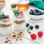 Protein-Packed Yogurt Bowl. Healthy eating: two glass containers with homemade yogurt and granola shot on breakfast table. Two spoons are beside the yogurt containers. Some berries complete the composition. The composition is at the left of an horizontal frame leaving useful copy space for text and/or logo at the right. Predominant color is green. High resolution 42Mp studio digital capture taken with Sony A7rii and Sony FE 90mm f2.8 macro G OSS lens
