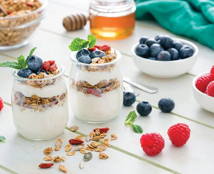 Protein-Packed Yogurt Bowl. Healthy eating: two glass containers with homemade yogurt and granola shot on breakfast table. Two spoons are beside the yogurt containers. Some berries complete the composition. The composition is at the left of an horizontal frame leaving useful copy space for text and/or logo at the right. Predominant color is green. High resolution 42Mp studio digital capture taken with Sony A7rii and Sony FE 90mm f2.8 macro G OSS lens