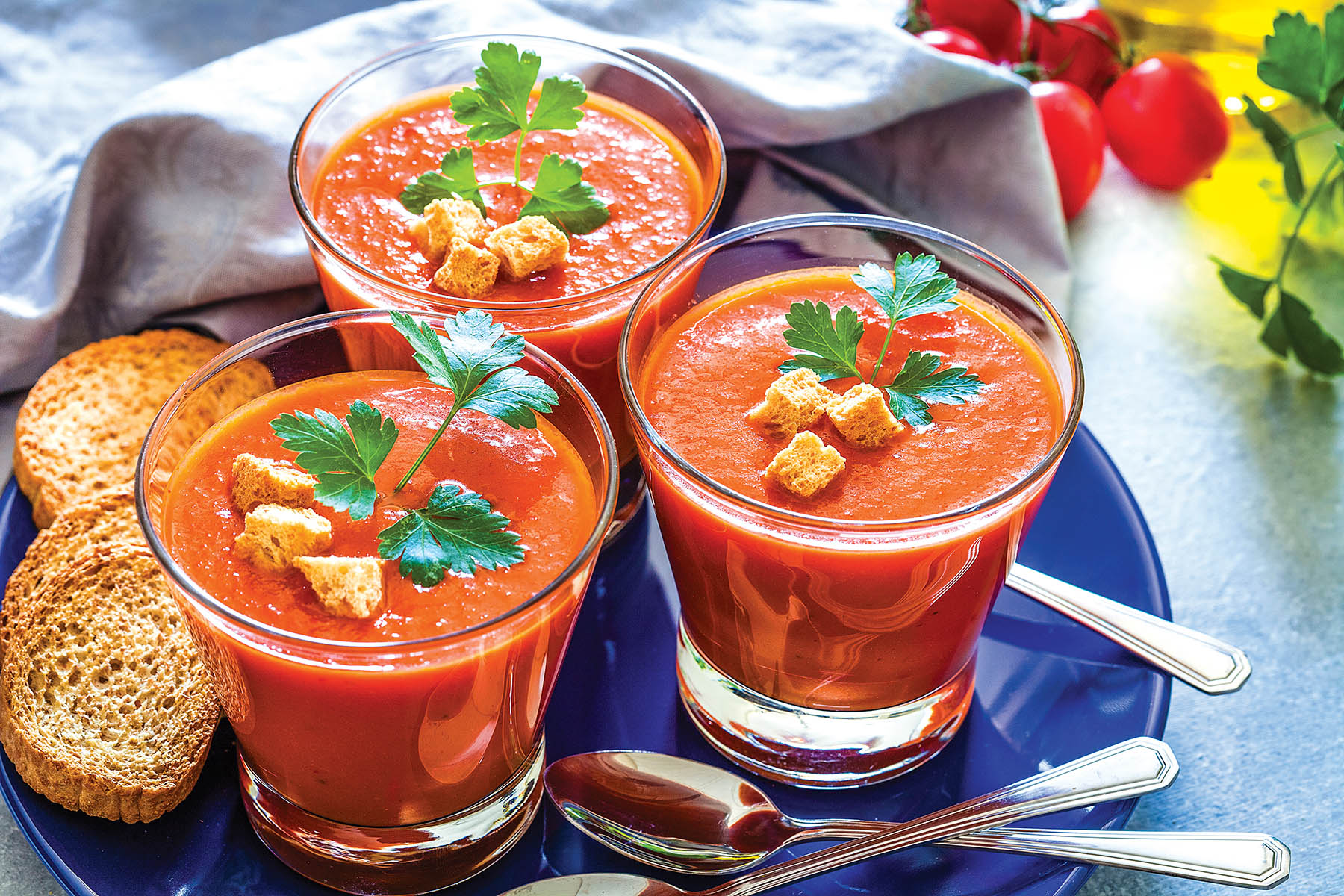 Three drinking glasses filled with Spanish gazpacho soup shot on bluish tint table. The gazpacho glasses are in a blue plate and some ingredients like tomatoes, parsley and olive oil complete the composition. Predominant colors are blue and red. High key DSRL studio photo taken with Canon EOS 5D Mk II and Canon EF 100mm f/2.8L Macro IS USM.