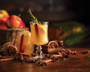 Mulled cider with slice apples, cinnamon, rosemary, and anise stars in glass cups on a brown rustic table. Holiday traditional hot drink. On a dark background kitchen utensils and burning candles.