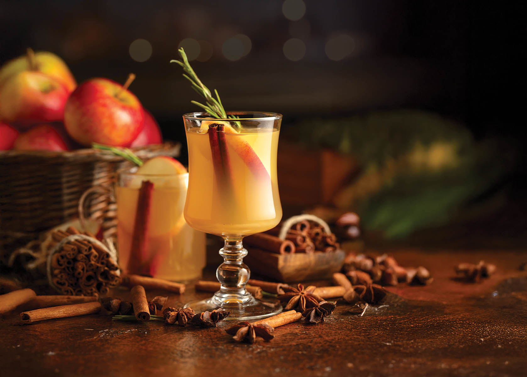 Mulled cider with slice apples, cinnamon, rosemary, and anise stars in glass cups on a brown rustic table. Holiday traditional hot drink. On a dark background kitchen utensils and burning candles.