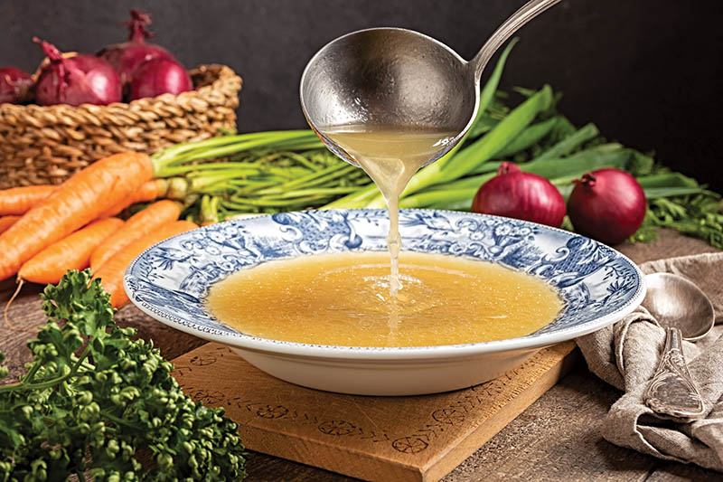 Pouring chicken bone broth from a ladle into a vintage plate, with fresh vegetables in the background