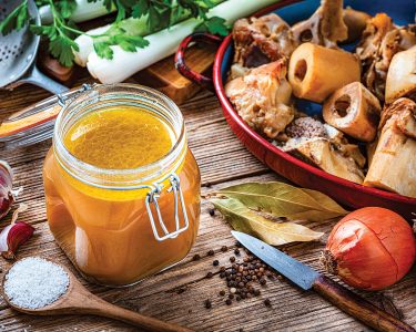 High angle view of healthy homemade bone broth in an open airtight glass container shot on rustic wooden table. Ingredients for cooking bone broth are all around the container. High resolution 42Mp studio digital capture taken with Sony A7rII and Sony FE 90mm f2.8 macro G OSS lens