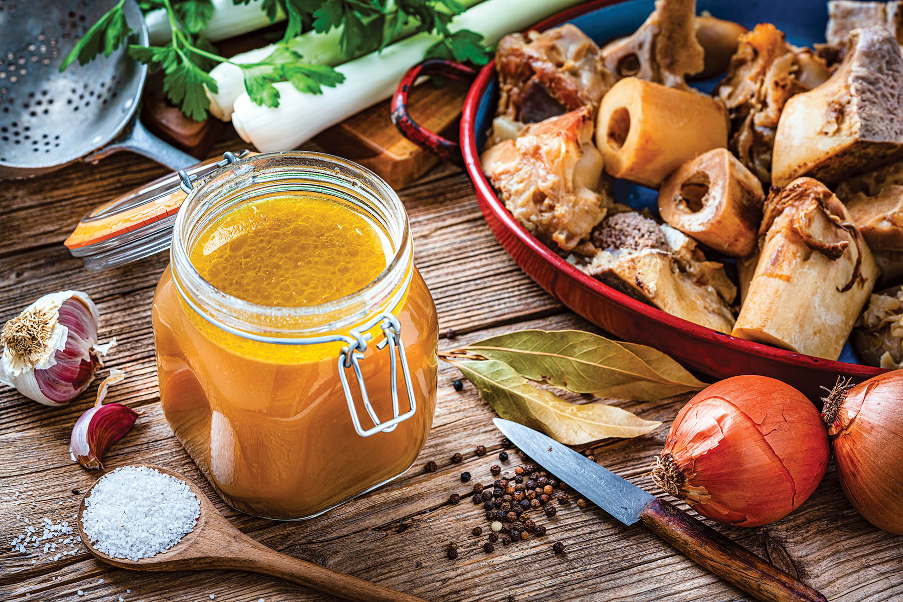 High angle view of healthy homemade bone broth in an open airtight glass container shot on rustic wooden table. Ingredients for cooking bone broth are all around the container. High resolution 42Mp studio digital capture taken with Sony A7rII and Sony FE 90mm f2.8 macro G OSS lens