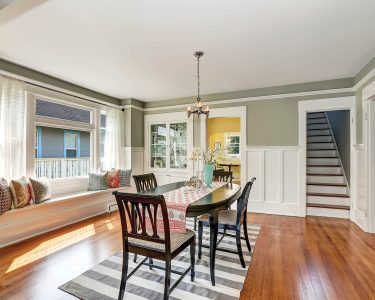 View of a classic dining room with gray walls and medium tone hardwood floors. Glass door cabinet and cozy window seat with pillows. Northwest, USA