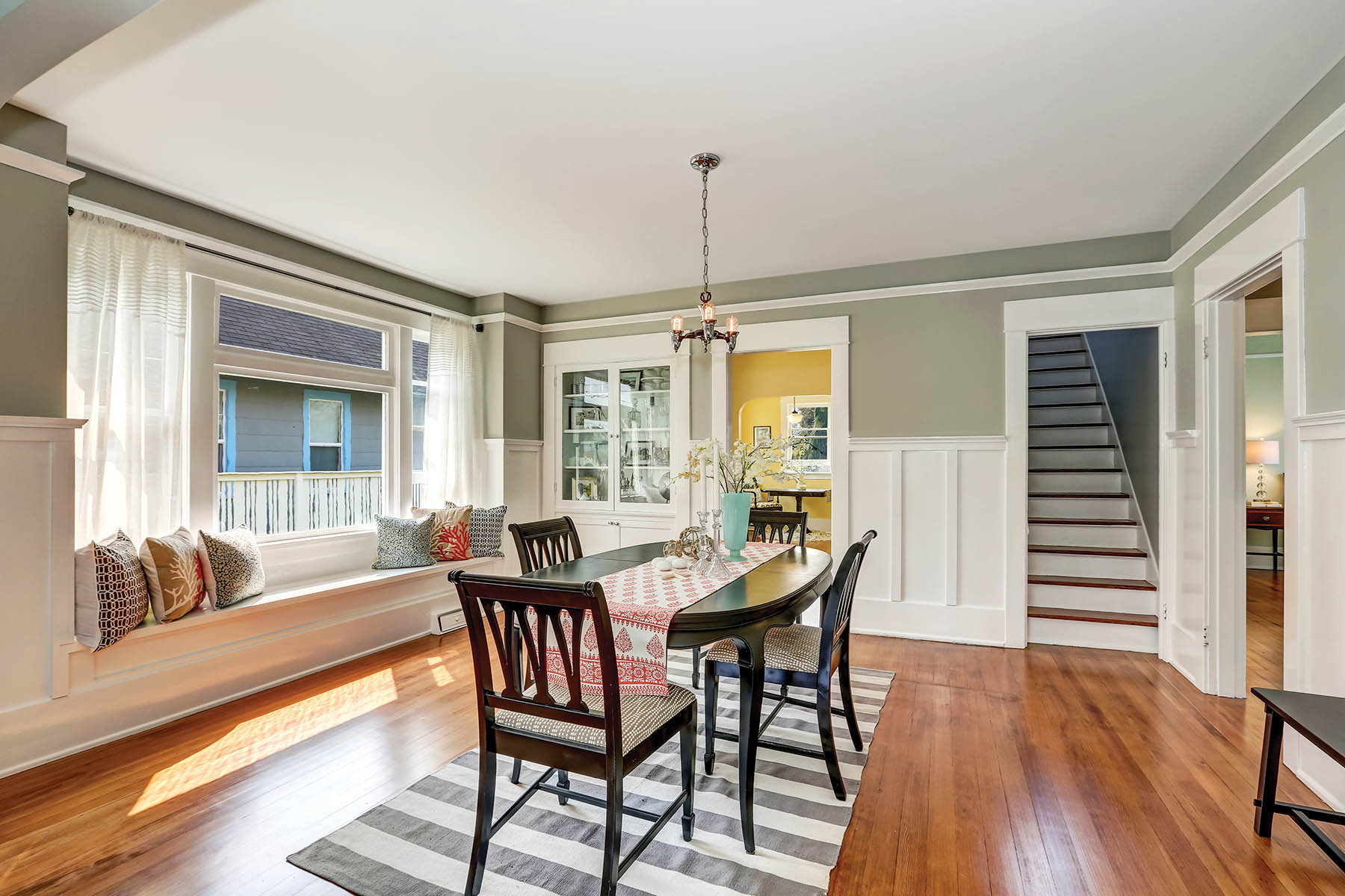 View of a classic dining room with gray walls and medium tone hardwood floors. Glass door cabinet and cozy window seat with pillows. Northwest, USA