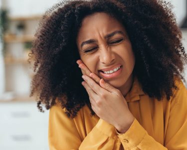 Young African American woman suffering from strong toothache touching her cheek because of acute pain at home