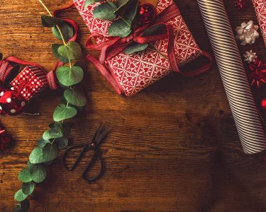 Woman packing contemporary christmas gifts, on table with red rustic paper and green leaves.
