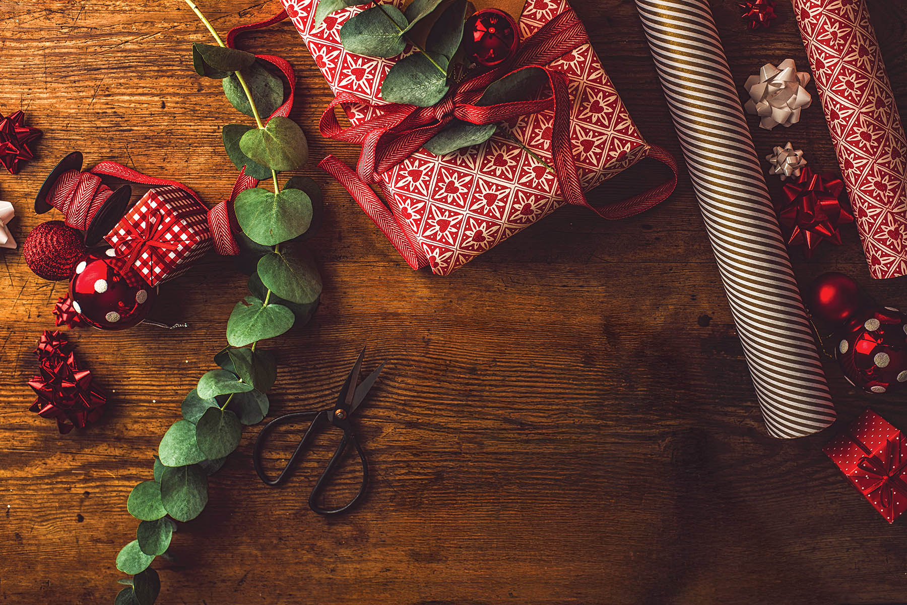 Woman packing contemporary christmas gifts, on table with red rustic paper and green leaves.