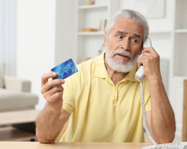 Confused senior man with credit card talking on phone at home. Be careful - fraud
