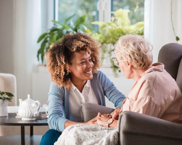 Female home caregiver talking with senior woman, sitting in living room and listening to her carefully.