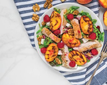 Grilled chicken fillet salad with grilled peaches, raspberries, walnuts and mixed greens on a plate with napkin and fork on white marble background. Top view. Healthy diet food. Summer dinner.