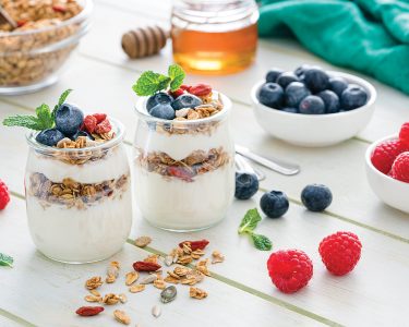 Protein-Packed Yogurt Bowl. Healthy eating: two glass containers with homemade yogurt and granola shot on breakfast table. Two spoons are beside the yogurt containers. Some berries complete the composition. The composition is at the left of an horizontal frame leaving useful copy space for text and/or logo at the right. Predominant color is green. High resolution 42Mp studio digital capture taken with Sony A7rii and Sony FE 90mm f2.8 macro G OSS lens