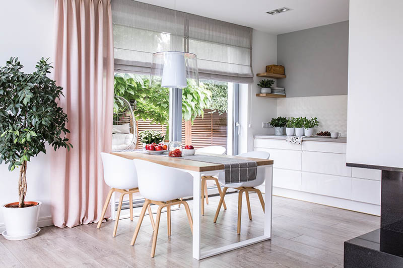 Gray roman shades and a pink curtain on big, glass windows in a modern kitchen and dining room interior with a wooden table and white chairs